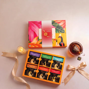 An open tea gift box displayed on a light beige surface, with its matching illustrated lid placed above it. The tea box contains multiple tea bags in colorful packets and is styled with a gold ribbon, a lit candle, and a glass cup of tea on a pale background.